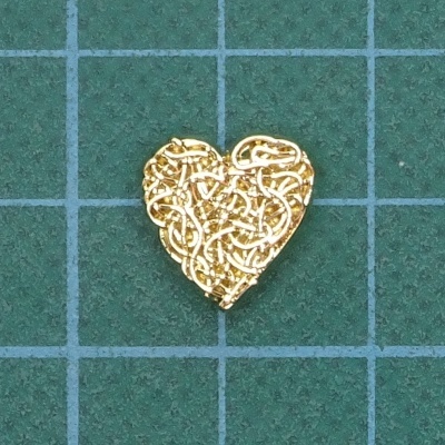 Peça dourada em forma de coração com padrão entrelaçado sobre fundo verde quadriculado