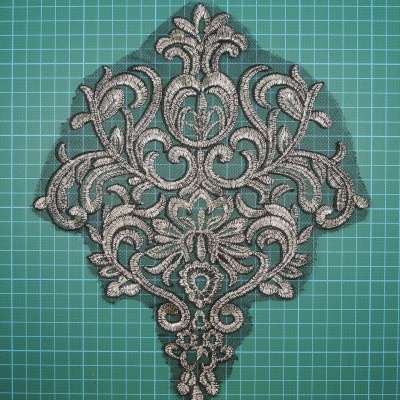 Aplicação de bordado ornamental bege em tecido sobre base quadriculada verde.