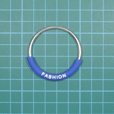 Anel metálico prateado com revestimento azul e texto FASHION sobre superfície quadriculada verde.