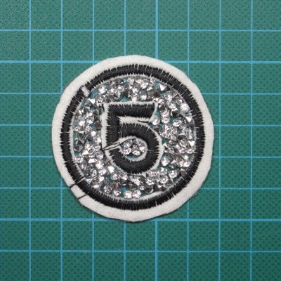 Emblema bordado com número 5 e brilhantes prateados sobre fundo verde quadriculado