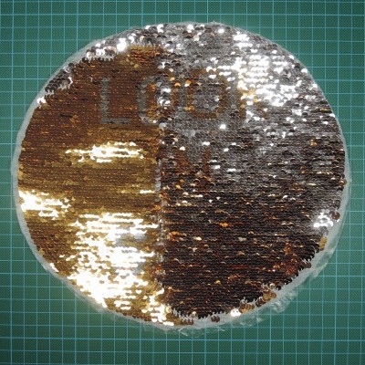 Almofada redonda com lantejoulas douradas e castanhas e palavra 'LOOK', sobre superfície verde quadriculada