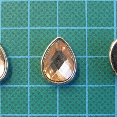 Três botões em forma de gota, dois com pedra âmbar facetada e moldura prateada, um mostrando o verso metálico, sobre fundo verde com linhas azuis.