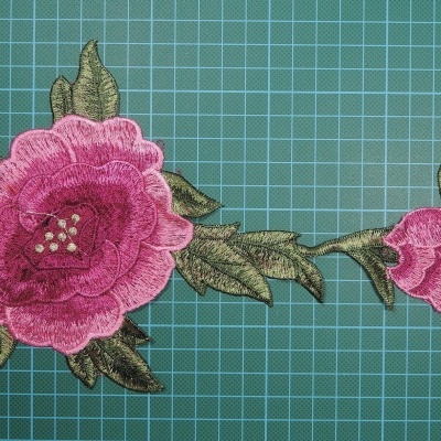 Aplicação bordada floral cor de rosa com folhas verdes sobre fundo verde quadriculado.