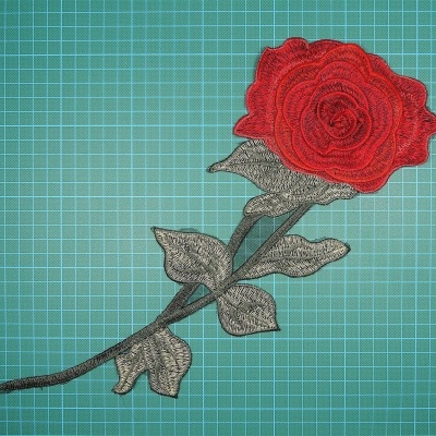 Aplicação bordada em forma de rosa vermelha com caule e folhas verdes sobre fundo verde quadriculado.