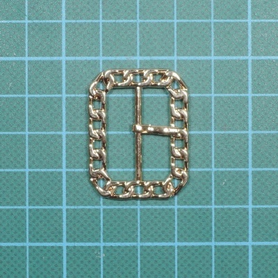 Fivela dourada retangular com padrão de elos sobre fundo quadriculado.