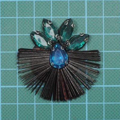 Broche com pedras azuis e verdes e franjas pretas sobre fundo verde quadriculado