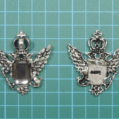 Dois emblemas metálicos prateados com detalhes de coroa, asas e perfil de rosto