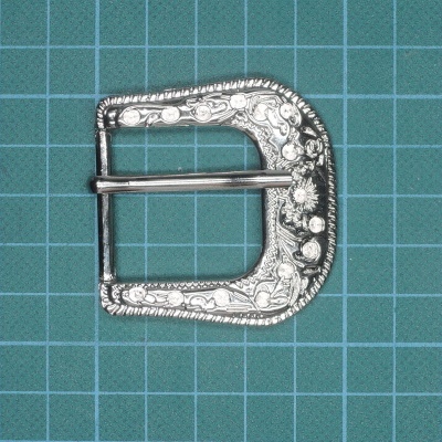 Fivela metálica prateada decorativa com padrão floral sobre superfície com grade