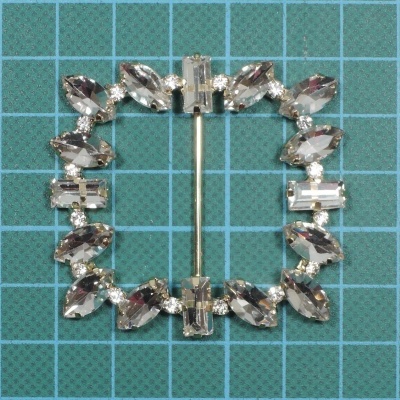 Broche quadrado com pedras transparentes em estrutura metálica prateada sobre fundo azul quadriculado