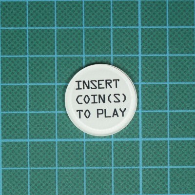 Botão branco com texto preto 'INSERT COIN(S) TO PLAY' sobre base verde de corte com grelha azul.