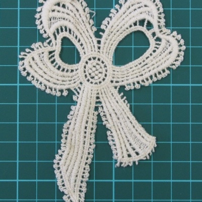 Aplique de renda branco em forma de laço sobre fundo com padrão de grade azul.