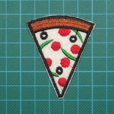 Peca de tecido bordado em forma de fatia de pizza sobre fundo verde quadriculado.