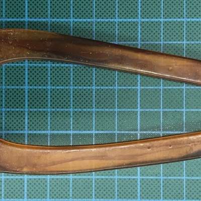 Objeto metálico bronze em forma de gota com recorte, sobre superfície azul quadriculada