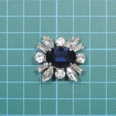 Broche com pedra azul central e pedras transparentes ao redor sobre fundo azul quadriculado