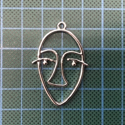 Pingente metálico prateado em forma de rosto abstrato sobre superfície quadriculada verde