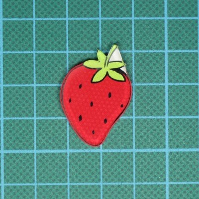 Patch de tecido com formato de morango vermelho e folhas verdes numa superfície verde quadriculada