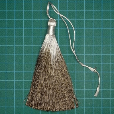 Pendente tassel de fios dourados e prateados com cordão fino, sobre fundo verde quadriculado