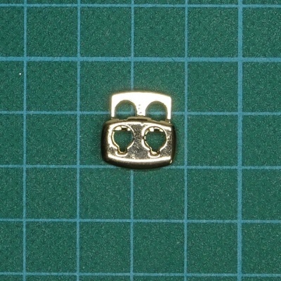 Peça metálica dourada com dois orifícios sobre superfície verde quadriculada