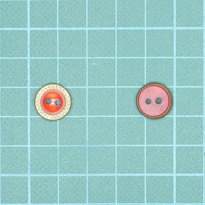 Dois botões redondos, um castanho claro com círculo laranja, outro rosa com rebordo vermelho, sobre fundo azul quadriculado