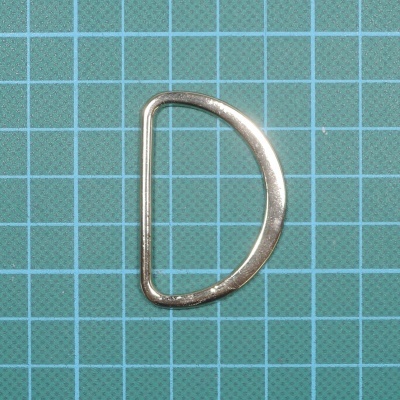 Anel metálico em forma de D prateado sobre base verde com grelha azul quadriculada