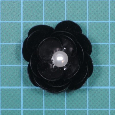 Broche preto em forma de flor com pérola branca no centro sobre fundo azul quadriculado