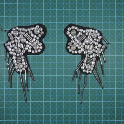 Par de aplicações decorativas pretas com brilhantes e fios metálicos prateados sobre superfície verde quadriculada