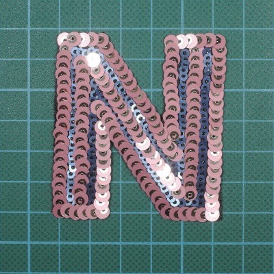 Aplicação letra N de lantejoulas cor-de-rosa e lilás sobre fundo de tecido verde com quadrícula branca