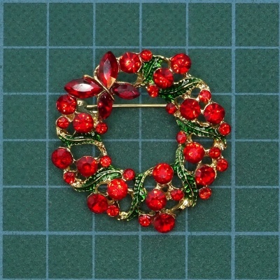 Broche dourado com pedras vermelhas e folhas verdes em fundo quadriculado