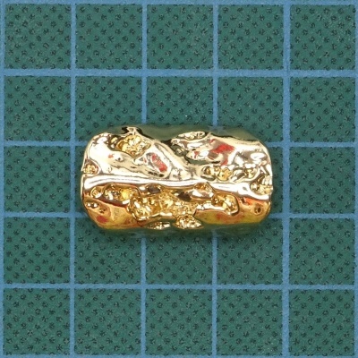 Peça metálica dourada com textura irregular sobre tabuleiro verde com quadrícula azul