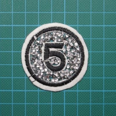 Emblema redondo branco com número 5 e cristais, sobre superfície de corte verde com grelha