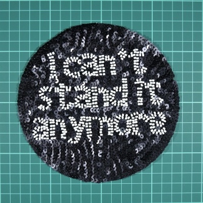 Patch redondo de lantejoulas pretas e brancas com texto em inglês sobre superfície verde com grelha.