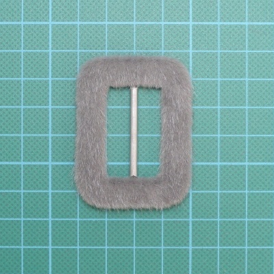 Fivela quadrada cinza com pelo e barra metálica central sobre superfície quadriculada verde