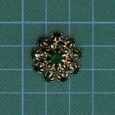 Botão decorativo redondo dourado com pedras verdes e transparentes em fundo quadriculado azul