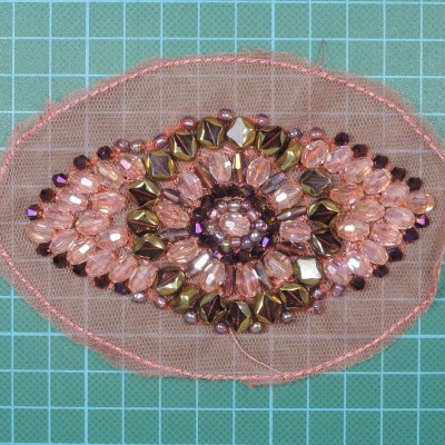 Aplicação decorativa em forma de olho com missangas cor de rosa e douradas em tela rosa sobre superfície verde quadriculada