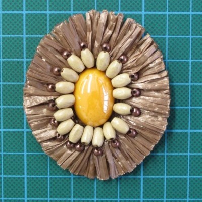 Broche oval com elemento central amarelo e contas decorativas, sobre fundo azul quadriculado