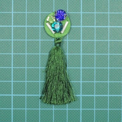 Brinco de franja verde com pedras coloridas sobre fundo de corte verde com quadrícula.