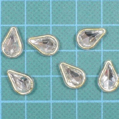 Seis pedras artificiais em forma de gota, transparentes com bordas douradas sobre superfície azul quadriculada