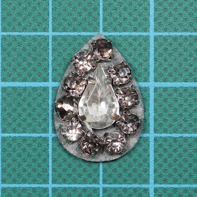 Broche gota com pedras transparentes e cinza sobre fundo metalizado