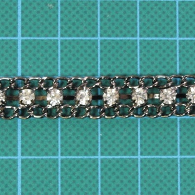 Tira decorativa com cristais transparentes em base metálica prateada sobre fundo de grade verde.