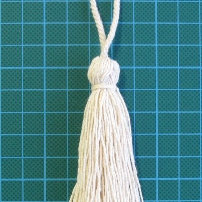 Pendente branco com franjas e laço sobre fundo verde quadriculado