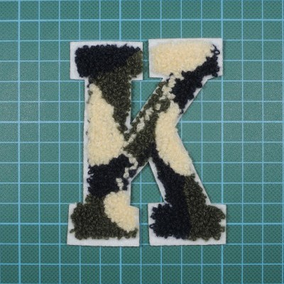 Patch felpudo letra K com padrão de cores preto, verde escuro e creme sobre superfície com padrão de grade verde.