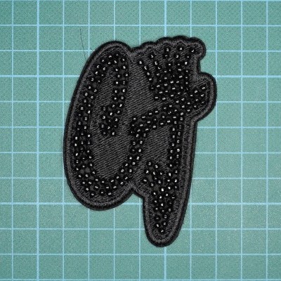 Patch bordado preto com letra G prateada sobre superfície de corte verde com grelha