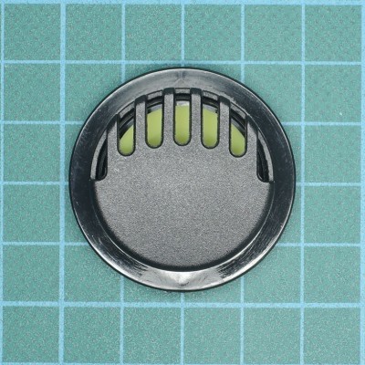 Tampa redonda preta com aberturas verticais e fundo verde em superfície de grade azul