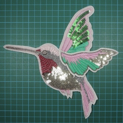 Adesivo bordado em forma de beija-flor com lantejoulas coloridas sobre superfície de corte verde