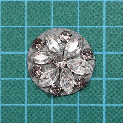 Botão decorativo redondo com pedras transparentes em padrão floral sobre base de feltro cinza