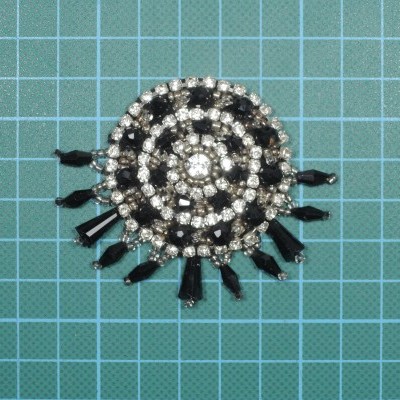 Broche redondo com pedras pretas e transparentes sobre base azul com grelha