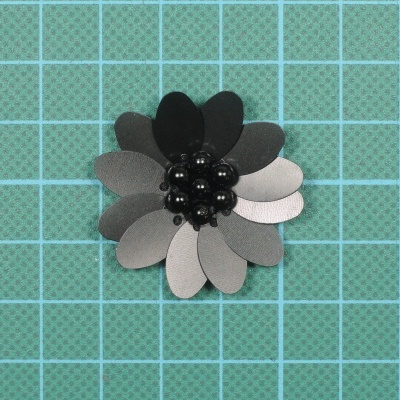 Broche flor cinza e preto com bolas no centro numa superfície quadriculada azul e verde