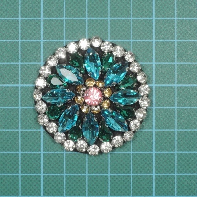 Broche redondo com pedras azuis, transparentes e rosa numa superfície quadriculada verde