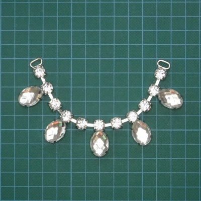 Bijuteria prateada com pedras facetadas ovais e quadradas sobre fundo quadriculado verde