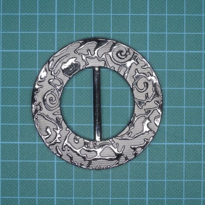Fivela metálica redonda com padrão gravado sobre superfície quadriculada verde